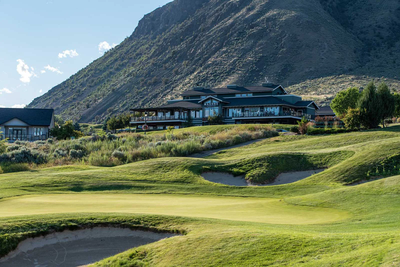 Golf course green and bunkers with clubhouse and patio set against Kamloops mountain landscape