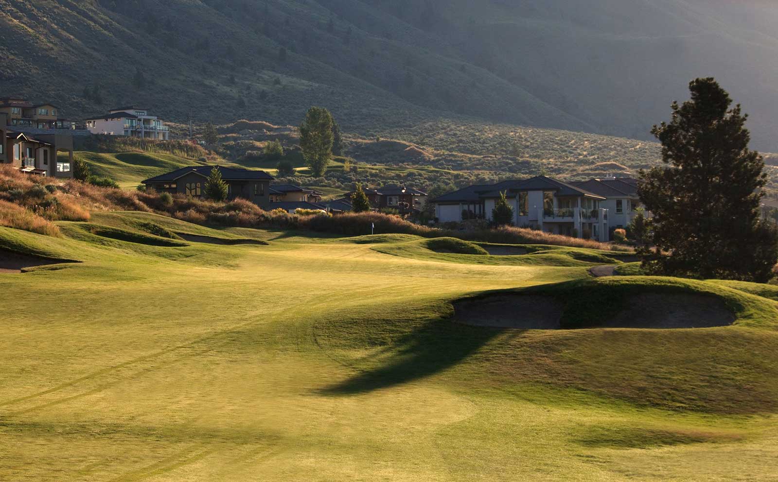 Sunlit golf fairway winding through hillside homes with bunkers and Kamloops valley backdrop
