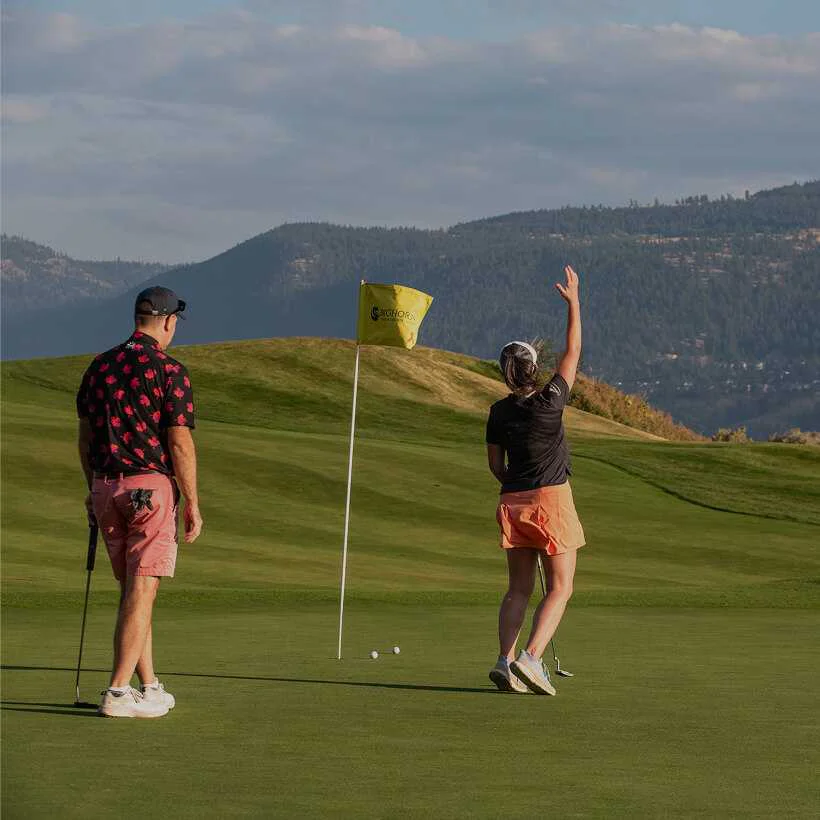 Two golfers on a green at Bighorn Golf & Country Club; one golfer raises her hand in celebration after a putt, while her partner watches, with rolling green hills and the Kamloops mountains in the background.