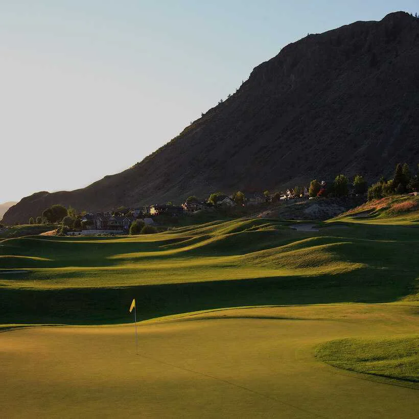 Sunset view of a golf green in the foreground with a yellow flag, looking toward rolling fairways and a steep mountain hillside under a clear sky in Kamloops.