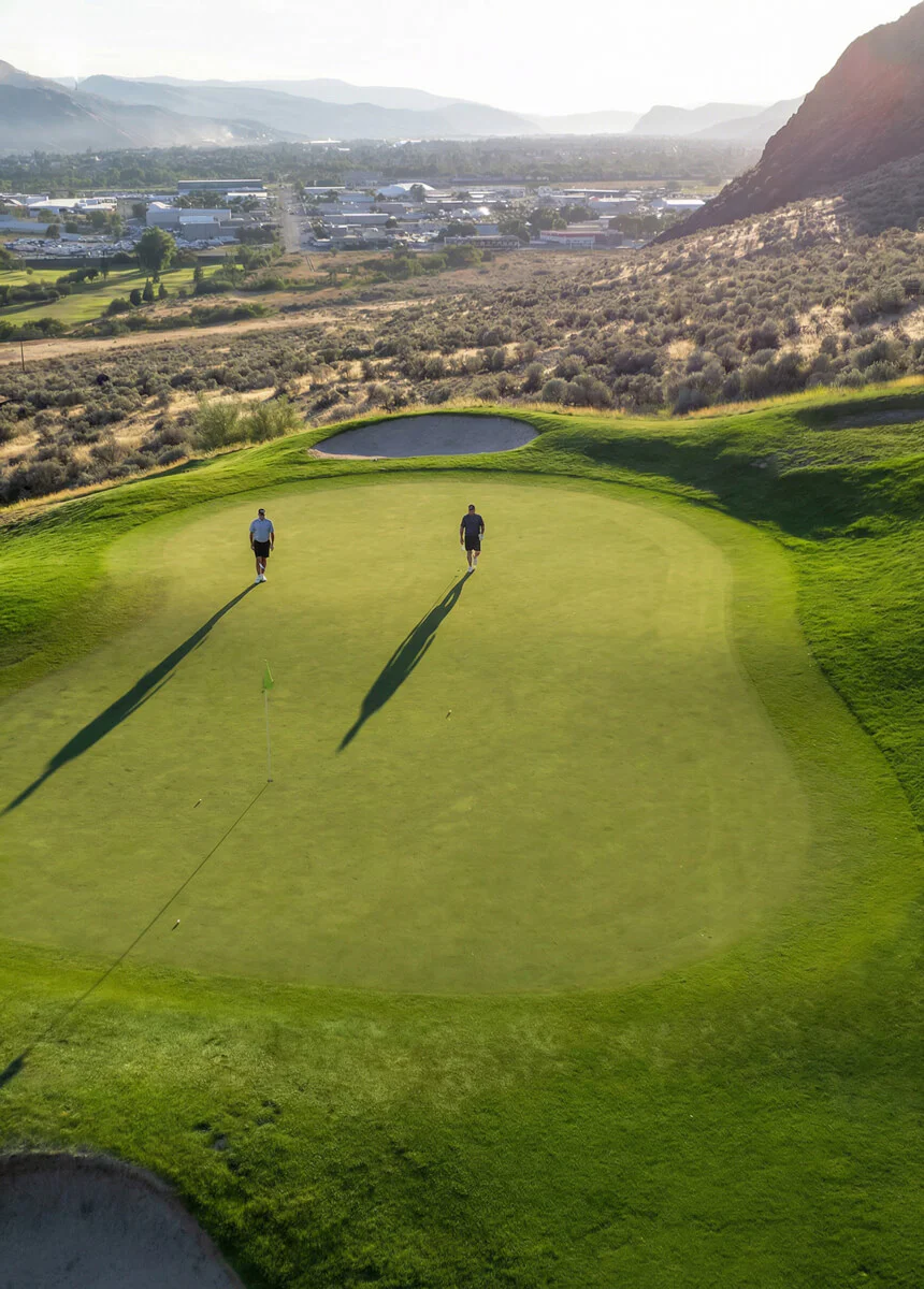 Aerial view of two golfers walking across a sunlit green at Bighorn Golf Course overlooking Kamloops valley